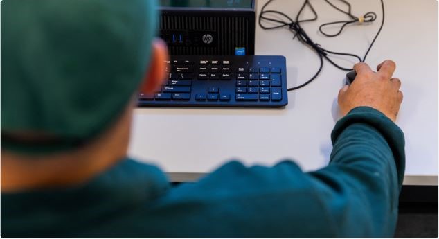 A man sitting infront of a desktop, holding the mouse. There is a keyboard too.