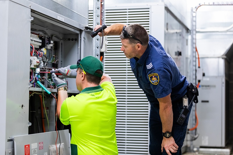 One man wearing a blue uniform, another wearing a high vis yellow shirt attending to an electrical  board