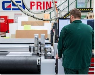 A man standing infront of a computer screen and printing something from a machine on his left